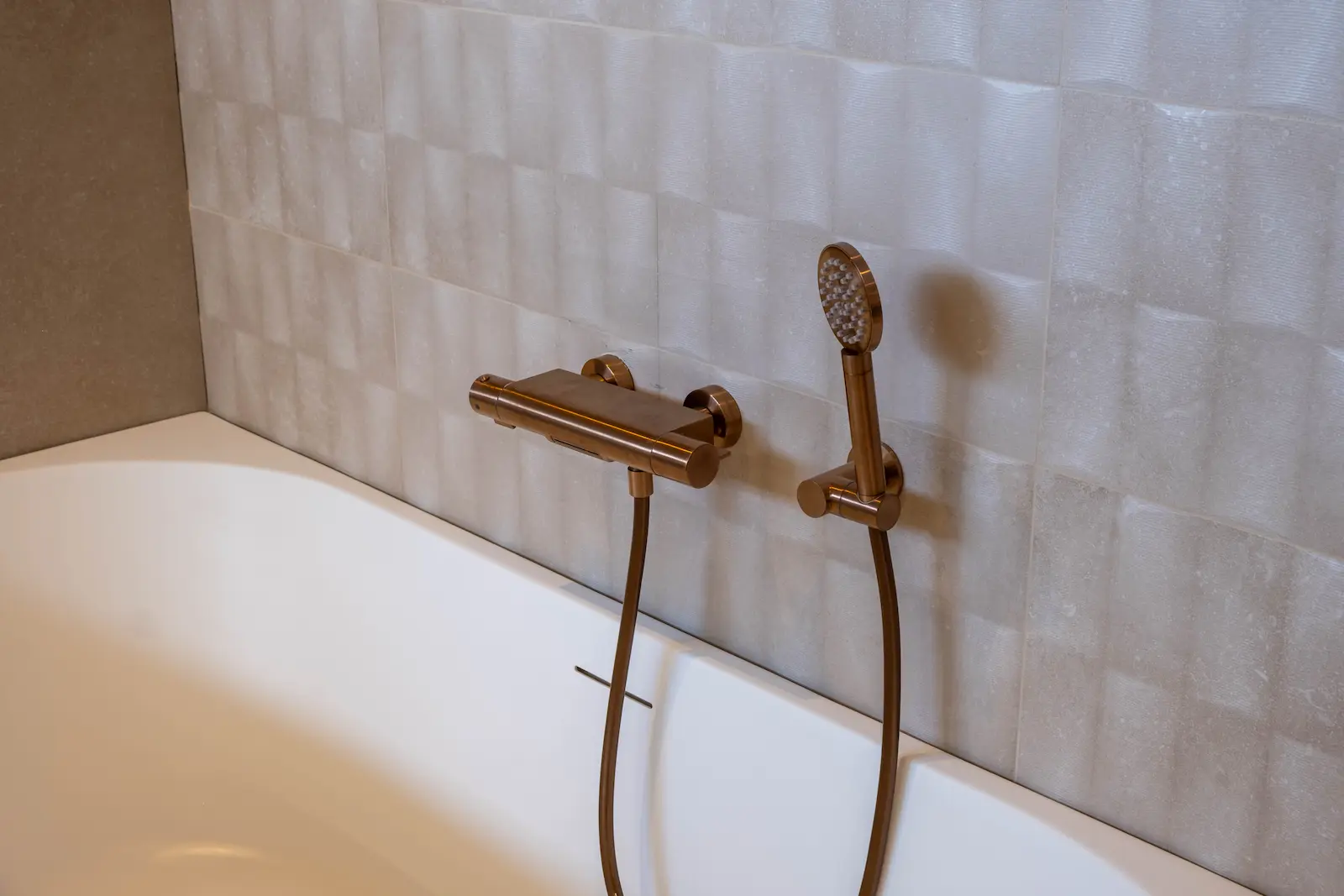 Thermostatische badkraan en handdouche in geborsteld koper bij een wit bad met reliëftegels