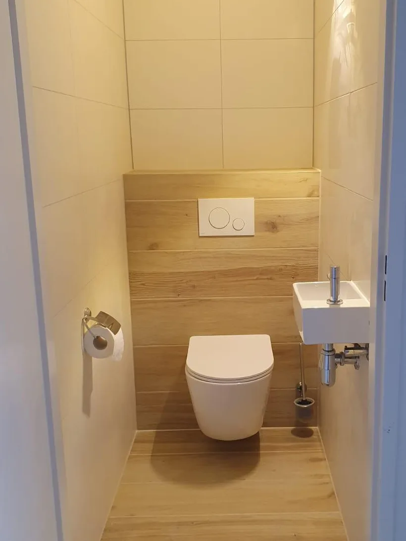 Gerenoveerd toilet met hangtoilet tegen een houtlook wand, houtlook vloertegels en een klein fonteintje met chroom kraan